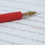 una penna rossa poggiata su un foglio di testo dove alcune parole sono riportate in rosso, l'immagine rappresenta il corso di editor e correttore di bozze