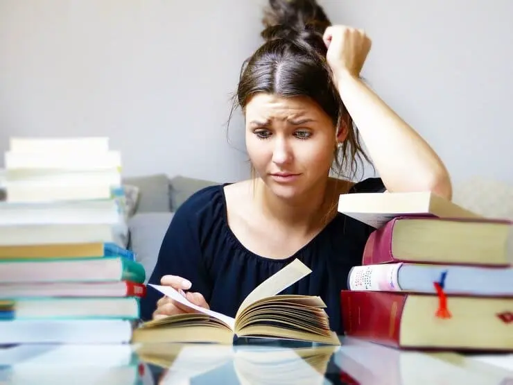una ragazza sconfortata che legge un libro circondata da pile di libri su una scrivania